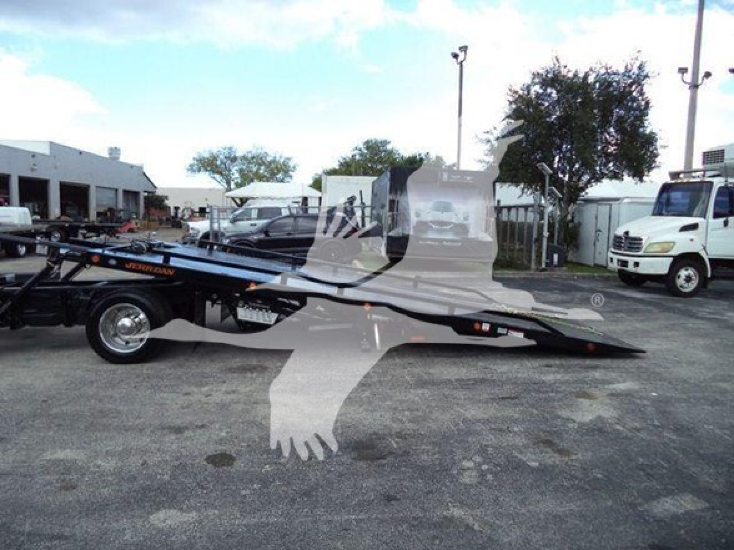 Flatbed tow truck chassis with rollback, Chevrolet Silverado 6500HD, image shows a blue/black flatbed with a white cab in background; no legible model text visible on the vehicle in the foreground.