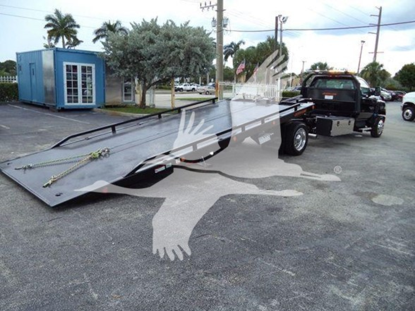 Truck with a long flatbed rollback trailer extending from the back of a black Chevrolet Silverado truck. There is a blue building and palm trees in the background.