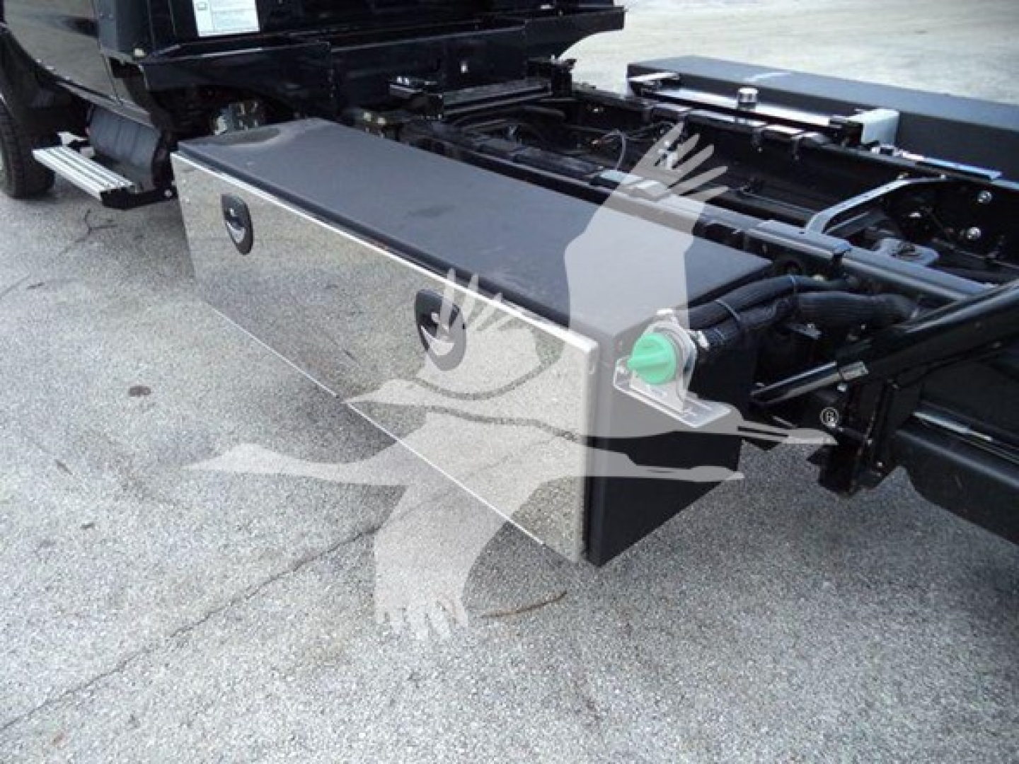 Triangle storage box mounted under truck chassis with a silver metallic toolbox showing two latches; green plug/valve and hydraulic lines nearby; part of rollback truck chassis visible.