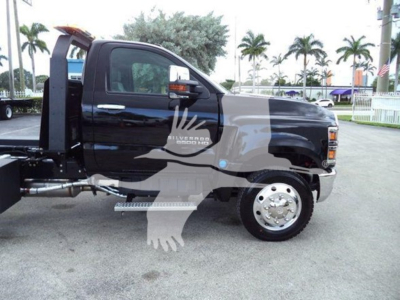 Truck side view showing a Chevrolet Silverado 6500HD rollback tow truck with black cab, chrome wheels, and equipment bed against a lot of palm trees.