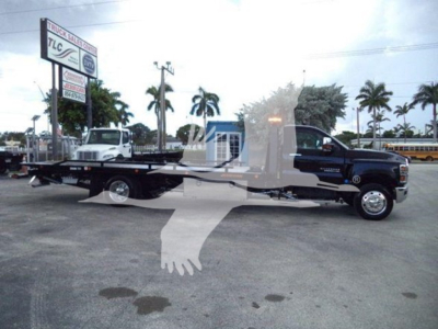Tow truck: Silverado 6500HD rollback with long flatbed on a dealership lot, palm trees in background, watermark overlay from dealership.