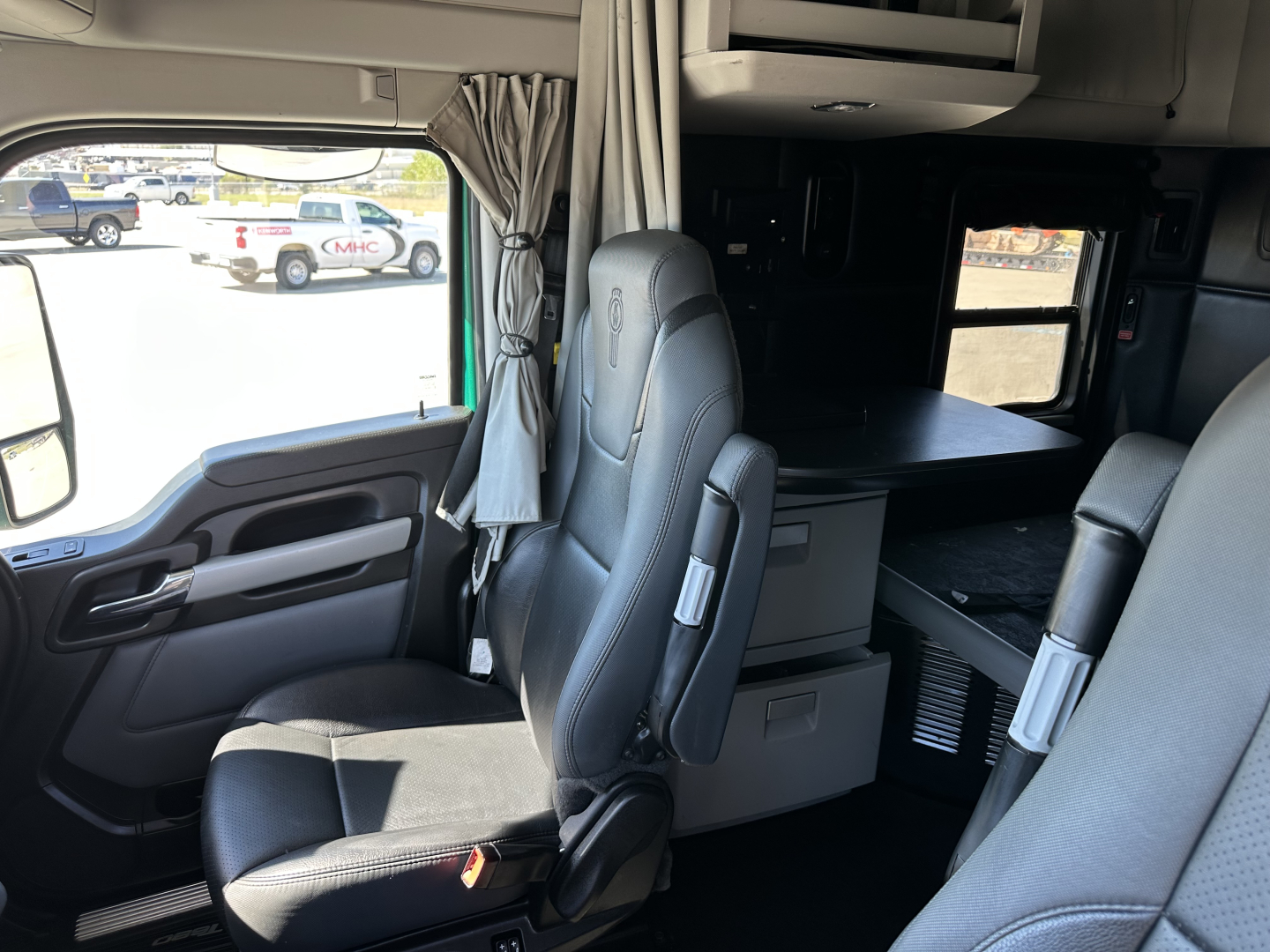 Kenworth T680 truck cab interior showing driver seat, dashboard, window, and sleeper area with curtains and storage cabinets.