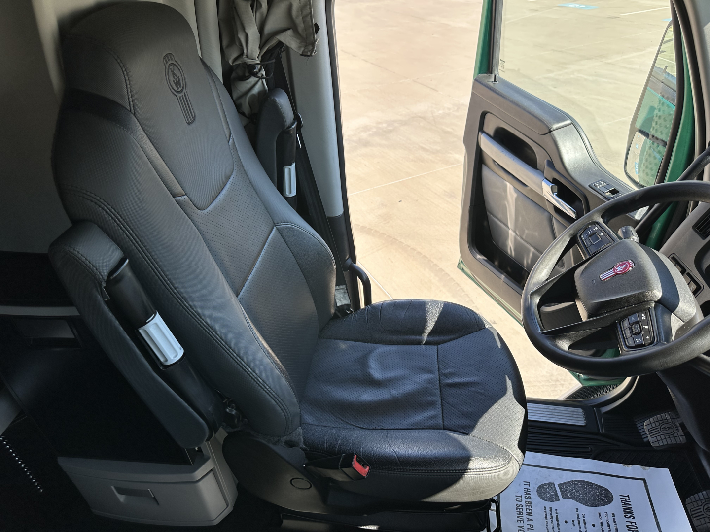 Interior of a 2023 Kenworth T680 cab showing driver seat and steering wheel, with door panel and window visible.