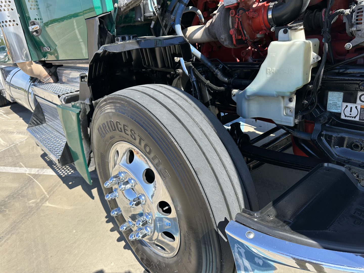 Kenworth T680 front wheel and engine bay view showing Bridgestone tire, chrome lug nuts, and part of the engine with coolant reservoir.