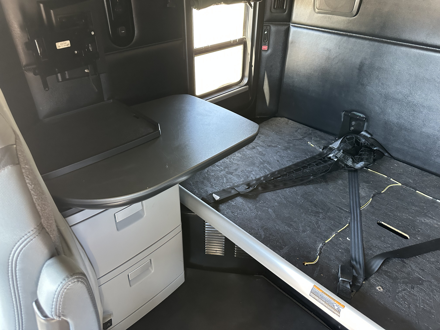 Interior of a 2023 Kenworth T680 sleeper cab showing a fold-out table, storage drawers, and seating area with seat belt; cab space and window visible.