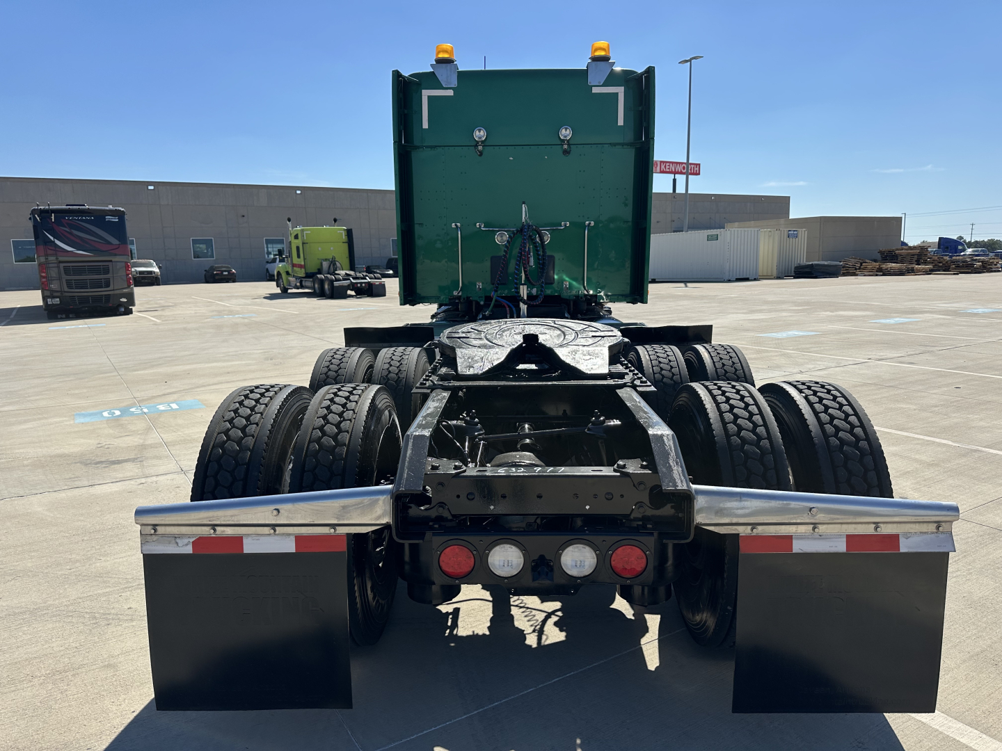 Kenworth T680 tractor rear view showing dual rear axles, fifth wheel, Mud flaps, and high-visibility lights on a sunny day.