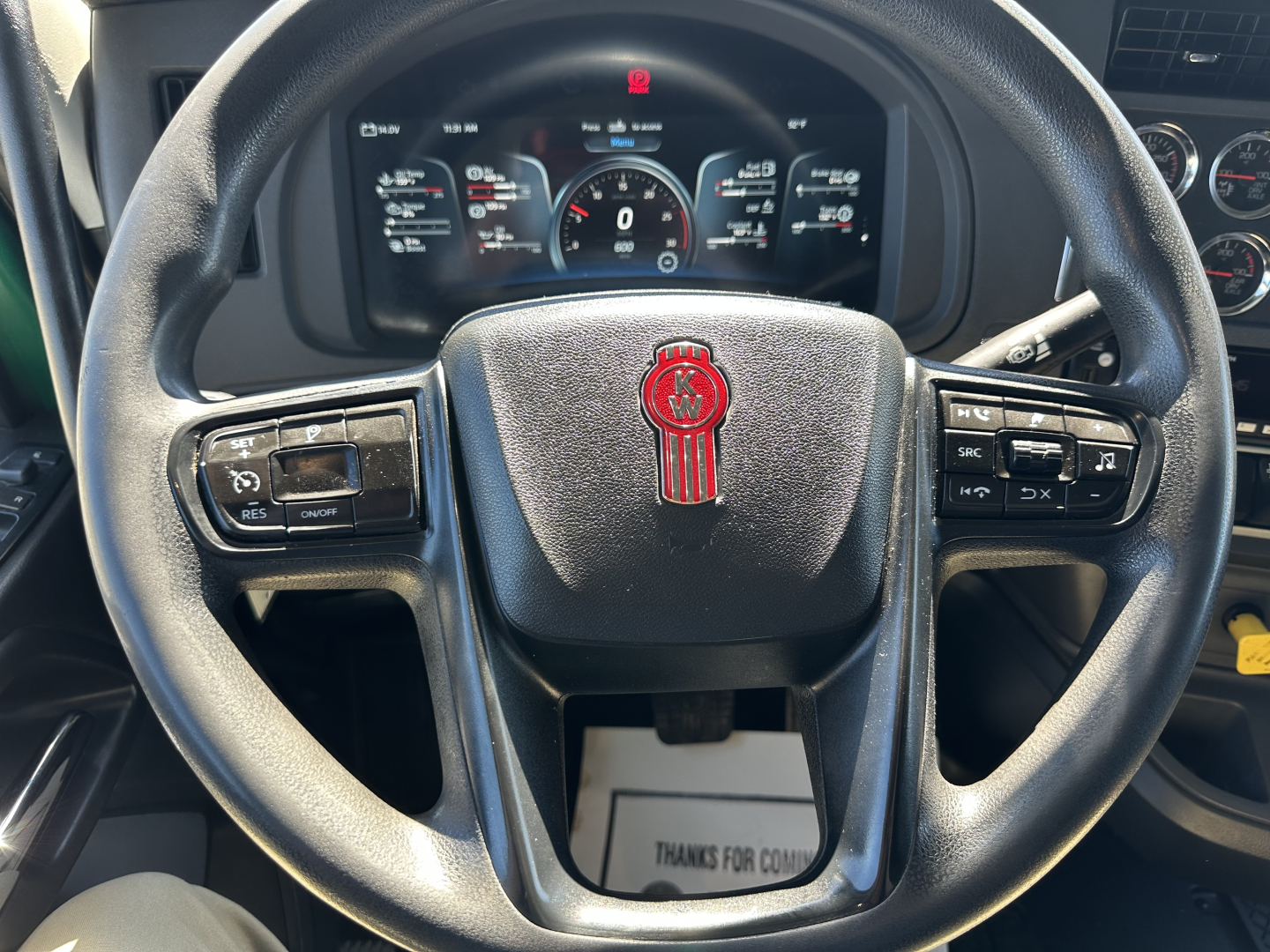 Steering wheel of a Kenworth truck with dashboard behind; Kenworth emblem centered, control buttons on both sides of the wheel, digital gauge cluster visible.