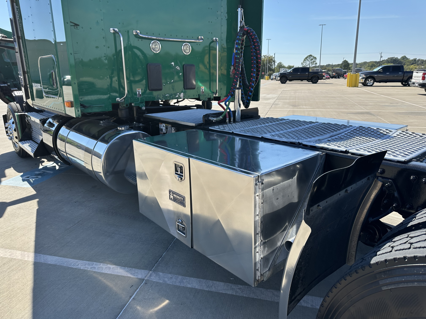 Kenworth T680 fuel tank and tool box area showing polished metal toolbox with two latches and brand emblem, fuel tank cylindrical behind, green cab, chrome steps.