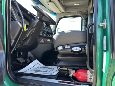 Kenworth T680 truck cab interior view from open driver door showing captain seat, dashboard, steering wheel, pedals, and a red fire extinguisher on the floor near the door. Paper along the floor mat.