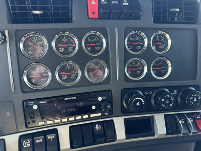 Kenworth T680 dash cluster with multiple gauges and a Kenworth radio in the center console. Various round gauges with red needles, climate controls on the right, hazard switch, and switch panels below.