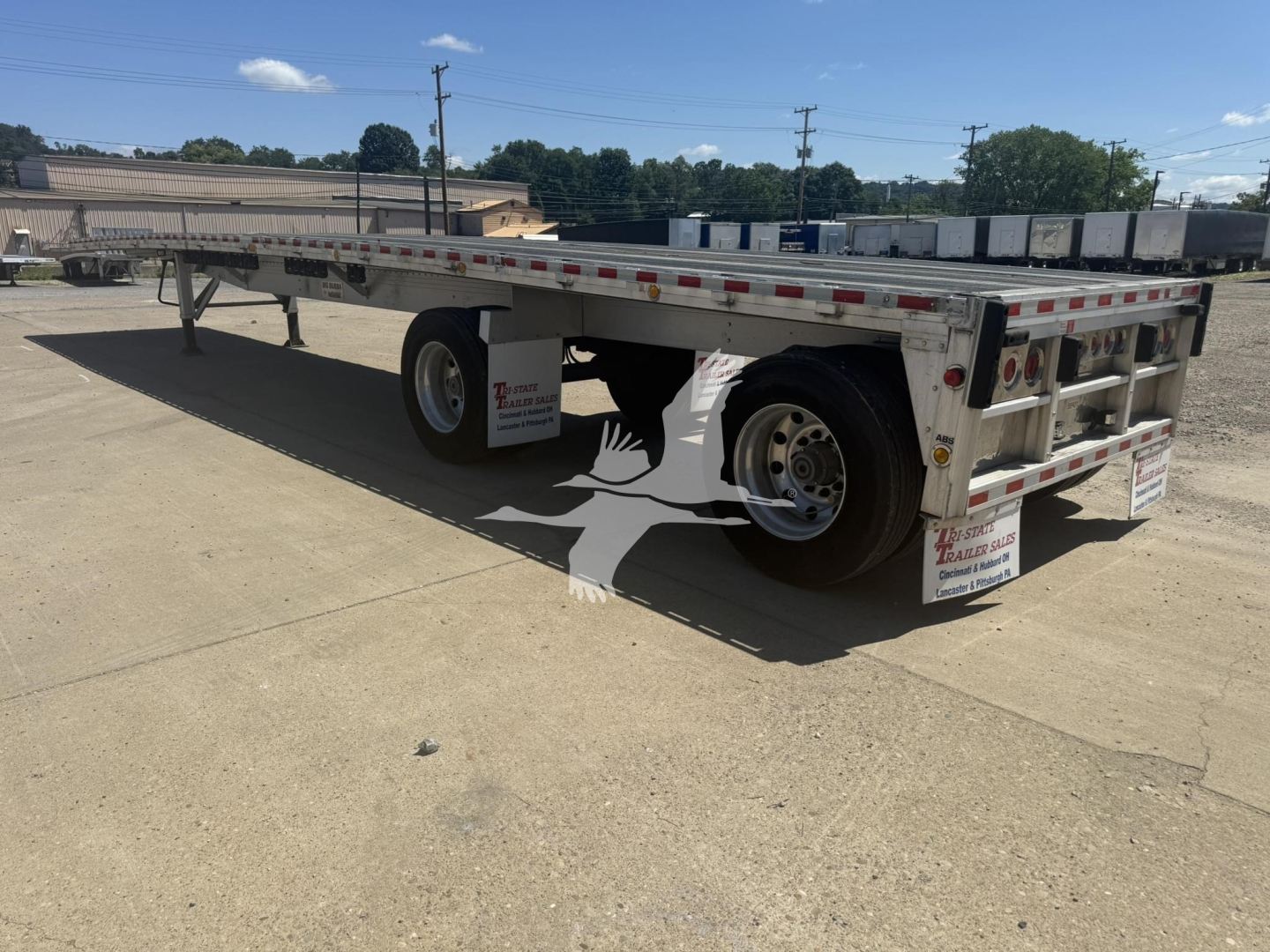 Flatbed trailer in a yard showing dual rear axles and reflective safety tape along the sides.