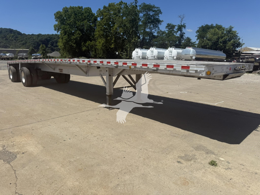 Flatbed trailer with dual rear axles and a long chrome fuel tank or cylindrical tanks on top, showing a Reitnouer Big Bubba trailer in a yard.