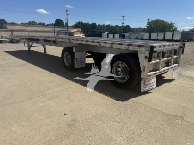 Flatbed trailer in a yard showing dual rear axles and reflective safety tape along the sides.