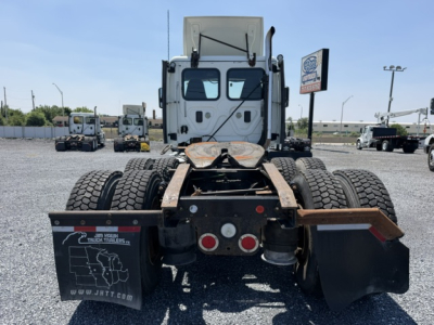 Rear view of a Freightliner Cascadia truck chassis showing dual rear axles, fifth wheel, and mud flaps with a dealership sign in the background.