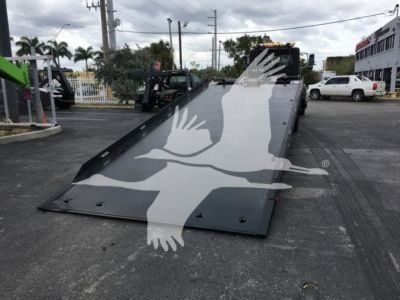 Tilting flatbed truck ramp on a rollback trailer with a Peterbilt 337 in background. Visible text includes a watermark with a bird logo.