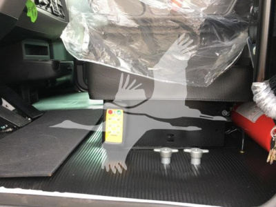 Interior of a 2022 Peterbilt 337 truck cab showing floor area with two small gray handles and a yellow control panel near the seat base, plastic seat cover, and partition in the background.