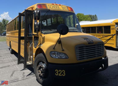School bus front-left view with yellow Freightliner C2 body, black bumper, number 323 on hood, and open entry doors visible.