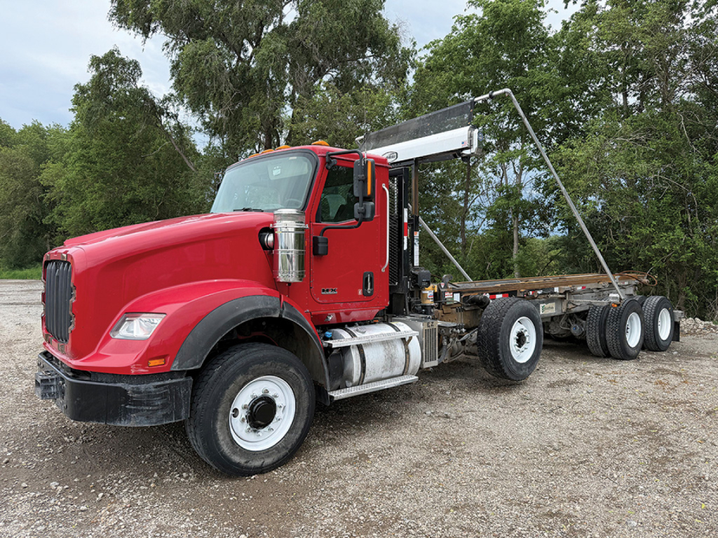 Internal combustion truck with a red cab and a long hydraulic rollback crane on a gravel lot, International HX620 model, front view.