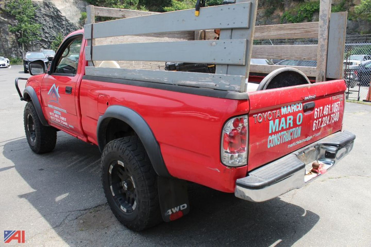 Rear-left view of red 2003 Toyota Tacoma pickup with wooden bed rails and 4x4 mud tires, tailgate and taillights visible, company graphics on doors and tailgate.