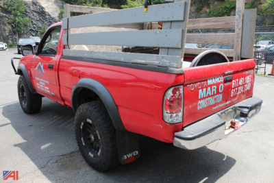 Rear-left view of red 2003 Toyota Tacoma pickup with wooden bed rails and 4x4 mud tires, tailgate and taillights visible, company graphics on doors and tailgate.