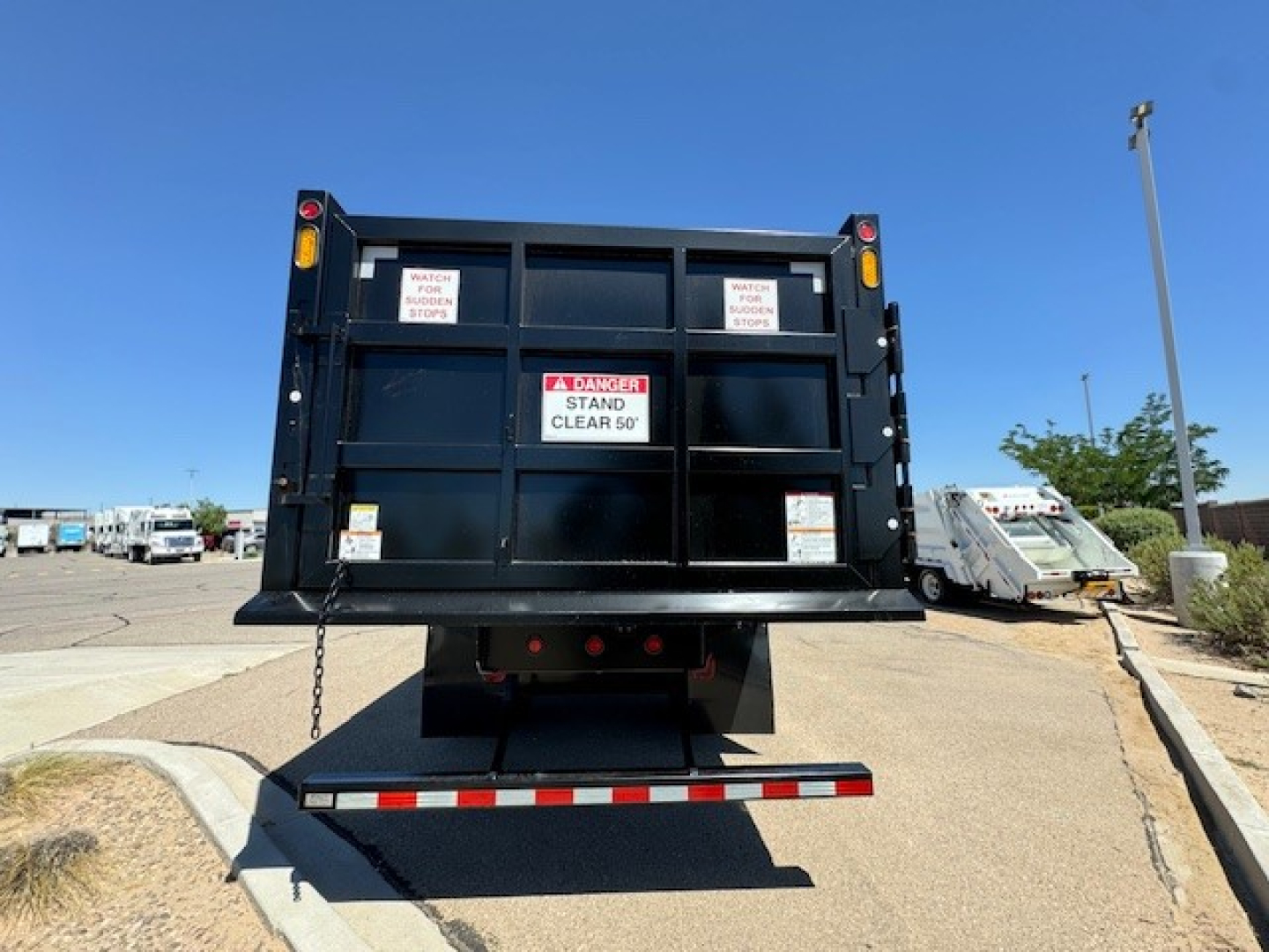 Rear view of a 2024 Hino L7 grapple truck bed with safety decals and warning labels on the tailgate, blue sky in background.