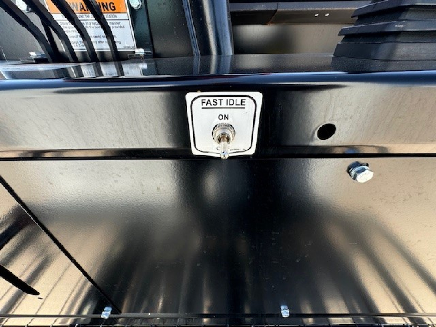 Fast idle switch with ON position on a metal surface of a truck component. Partial view of surrounding black metal panels and a small bolt.