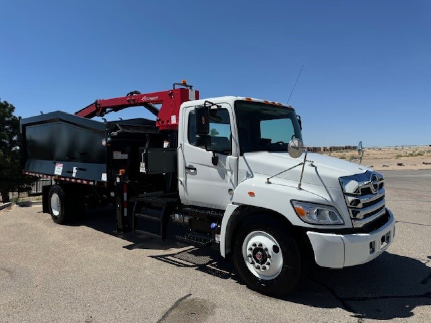 2024 Hino L7 grapple truck with crane and dump bed, white cab, red crane, black dump body. Visible text on truck includes none legible from distance.