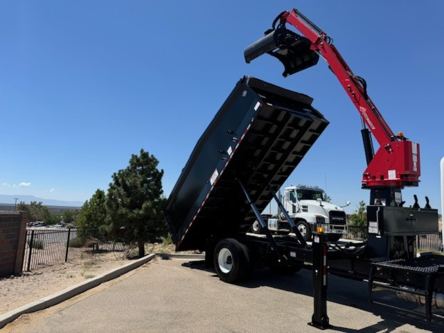 Truck with a raised dump bed and a red hydraulic grapple arm on a tilt bed in a parking lot.