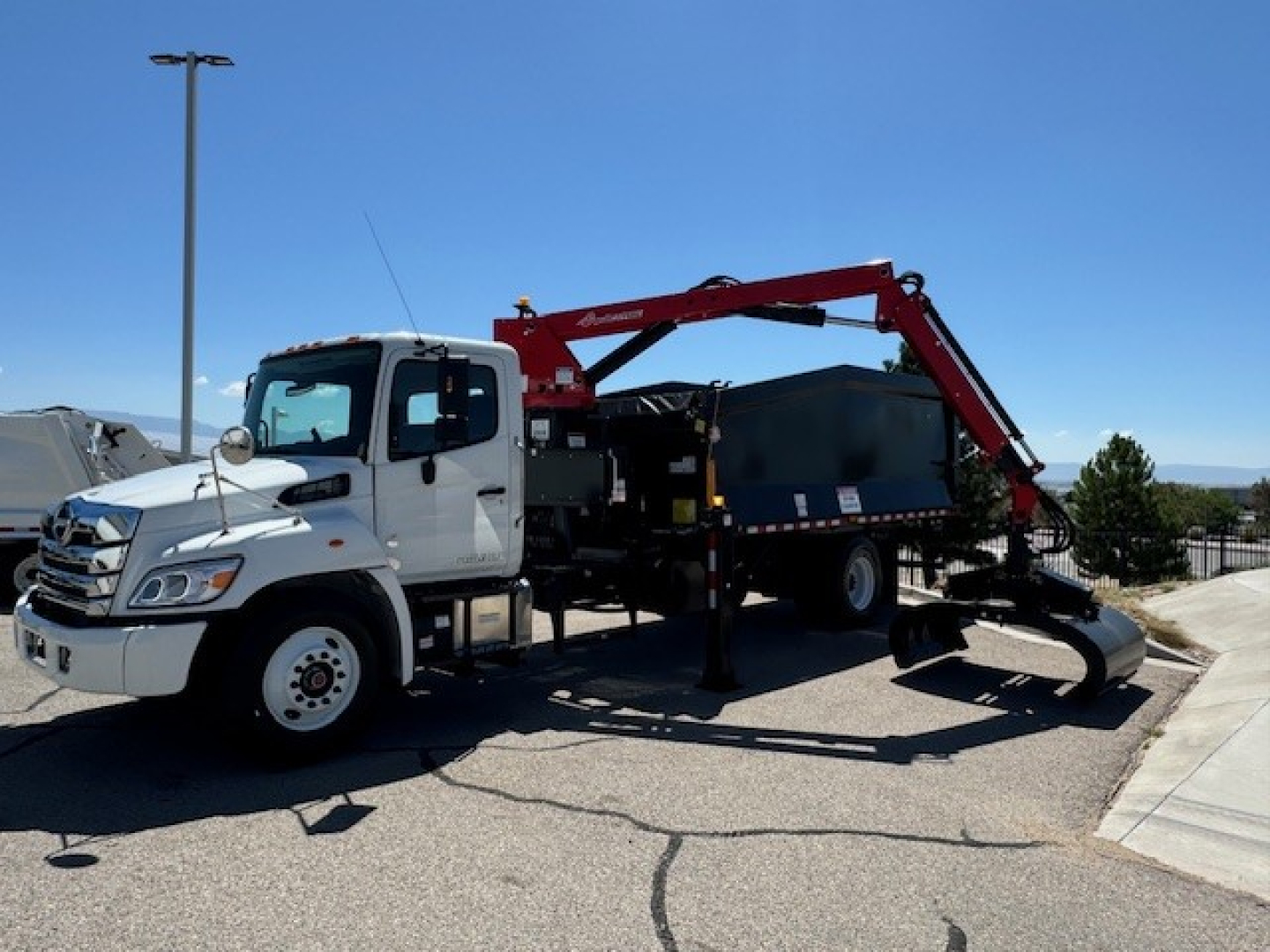 White Hino truck with a red hydraulic grapple arm and a mounted work bucket on a trailer, parked outdoors; no legible labels or stickers visible on the equipment.