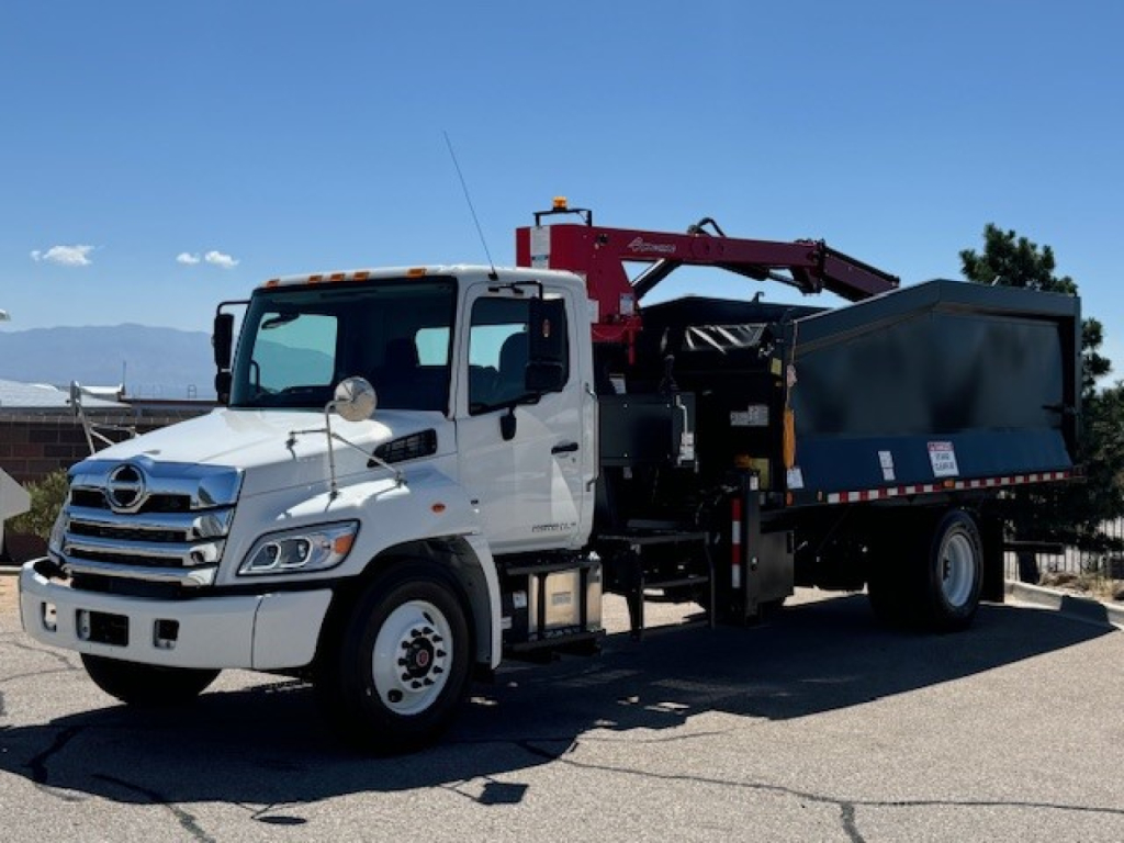 White 2024 Hino L7 grapple truck with a red hydraulic arm, dumping hopper on a gravel lot; visible front grille, cab, side mirrors, and black dump bed.