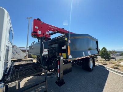 Grapple truck with a red Palfinger crane mounted on a blue dump body, in-service stabilizer legs deployed, side railings, and wheeled rear axle visible.