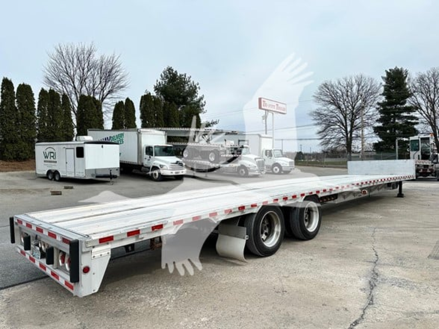 Flatbed trailer showing drop deck design with dual rear axles and aluminum deck. Side cargo protection and rear lights visible.