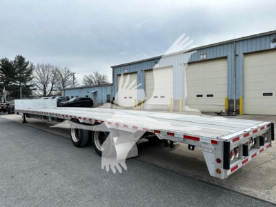 Flatbed trailer showing long aluminum deck and dual rear axles with drop deck style rear overhang. Side rails with reflective tape and rear lighting visible.