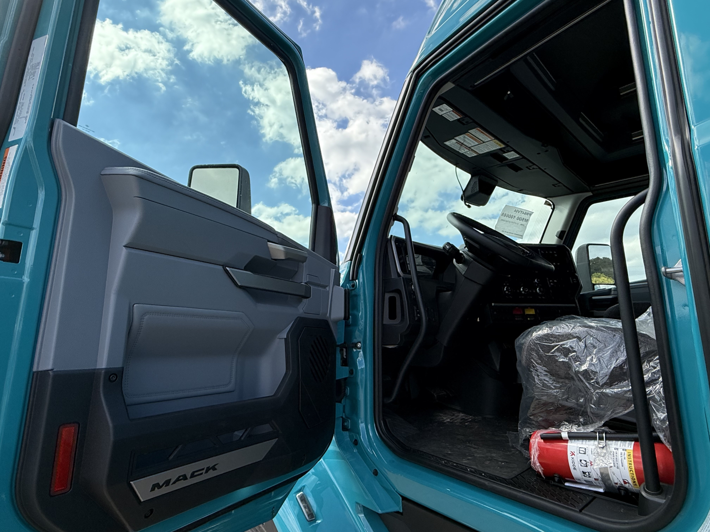 Truck cab interior with open door showing Mack branding on door panel and plastic-covered seat; fire extinguisher visible on passenger side floor; dashboard and steering wheel in view.