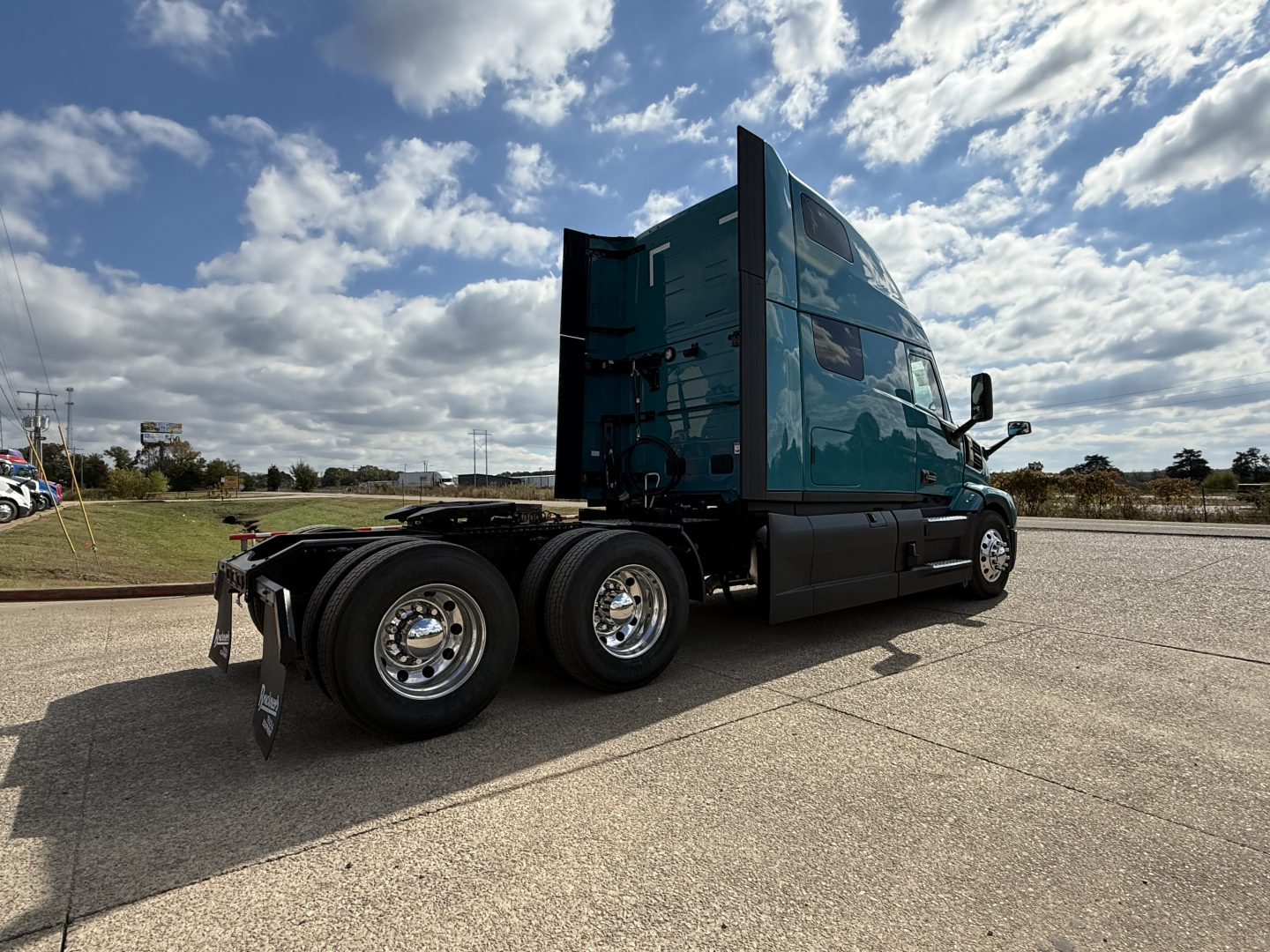 2026 Mack PR64T76H conventional sleeper truck with teal cab seen from rear-side, showing dual rear axles, chrome wheels, and the flatbed area behind the cab.