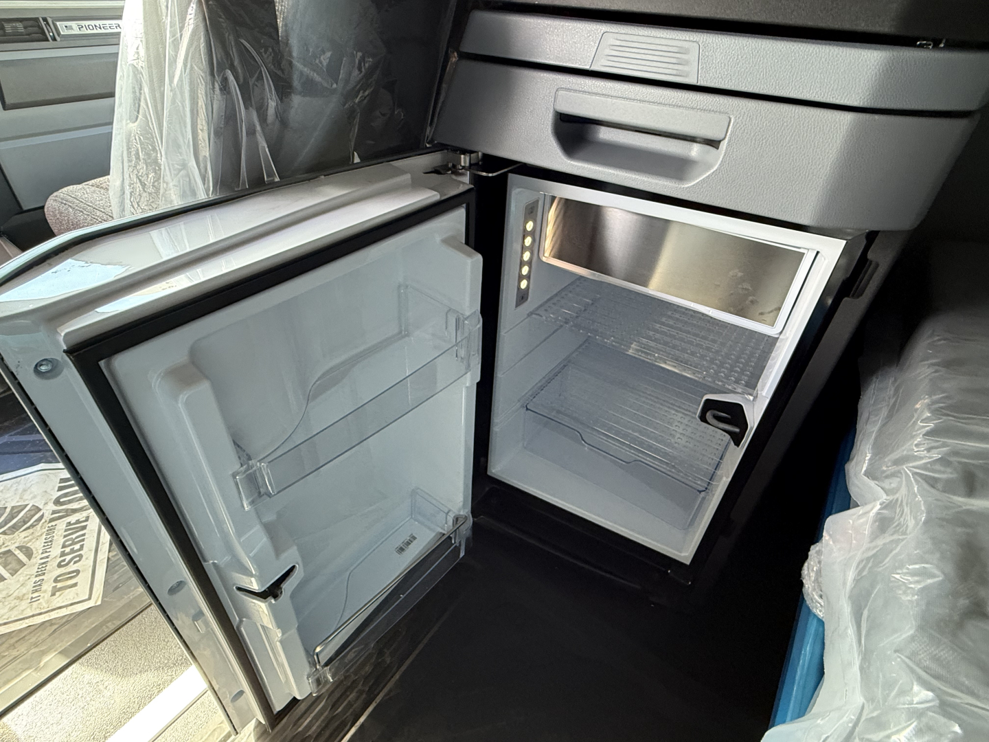 Interior of a Mack PR64T76H showing a small under-cabinet refrigerator/freezer with door open and empty shelves.
