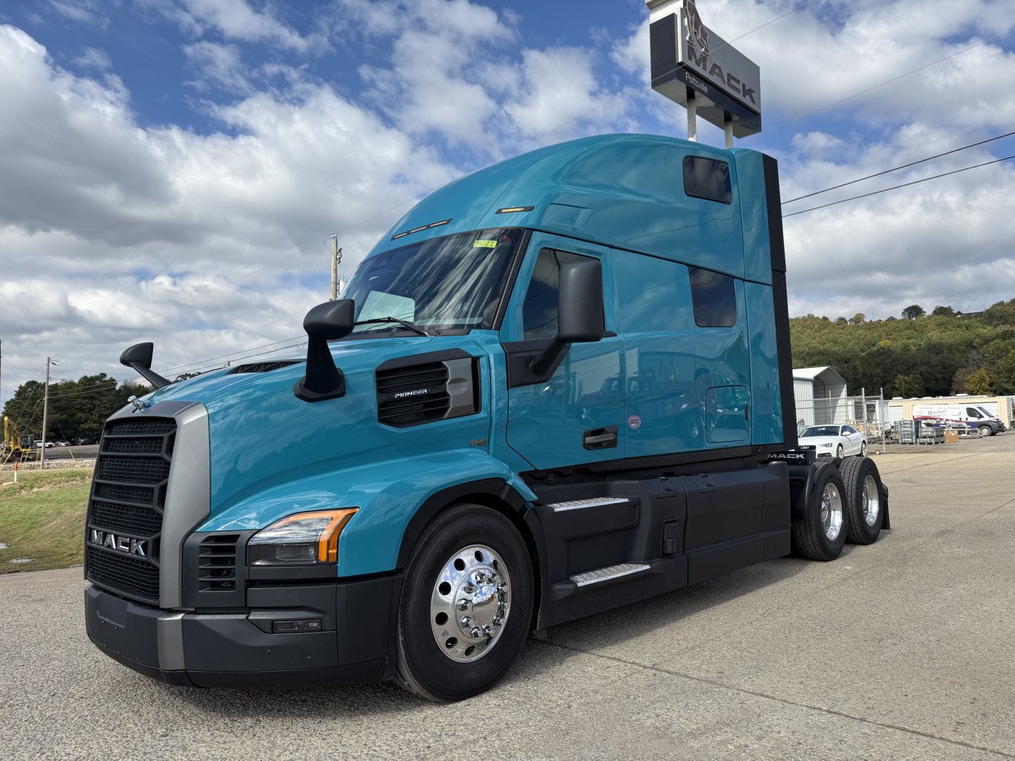 Blue Mack truck in a dealership lot with sleeper cab, showing front grille with Mack logo and side mirrors, no visible text other than the brand.