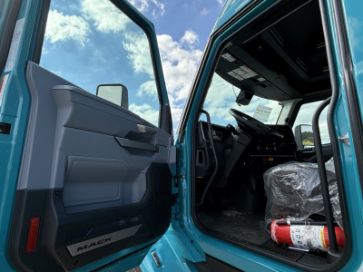 Truck cab interior with open door showing Mack branding on door panel and plastic-covered seat; fire extinguisher visible on passenger side floor; dashboard and steering wheel in view.