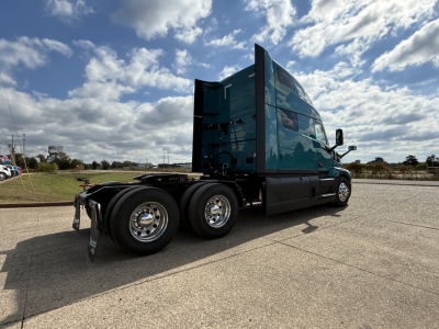 2026 Mack PR64T76H conventional sleeper truck with teal cab seen from rear-side, showing dual rear axles, chrome wheels, and the flatbed area behind the cab.