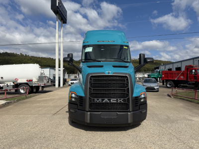 Front view of a teal 2026 Mack PR64T76H tractor with large black grille and MACK lettering, in a lot with other trucks and a blue sky.