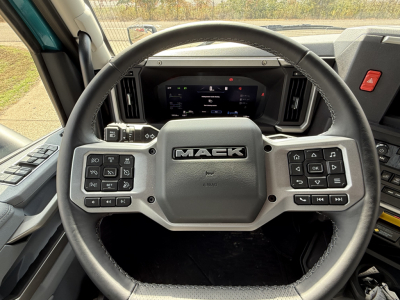 Mack PR64T76H truck interior steering wheel with dashboard and control panels. Visible text includes MACK on the center of the steering wheel, various control icons on both sides of the wheel, and an instrument display behind.