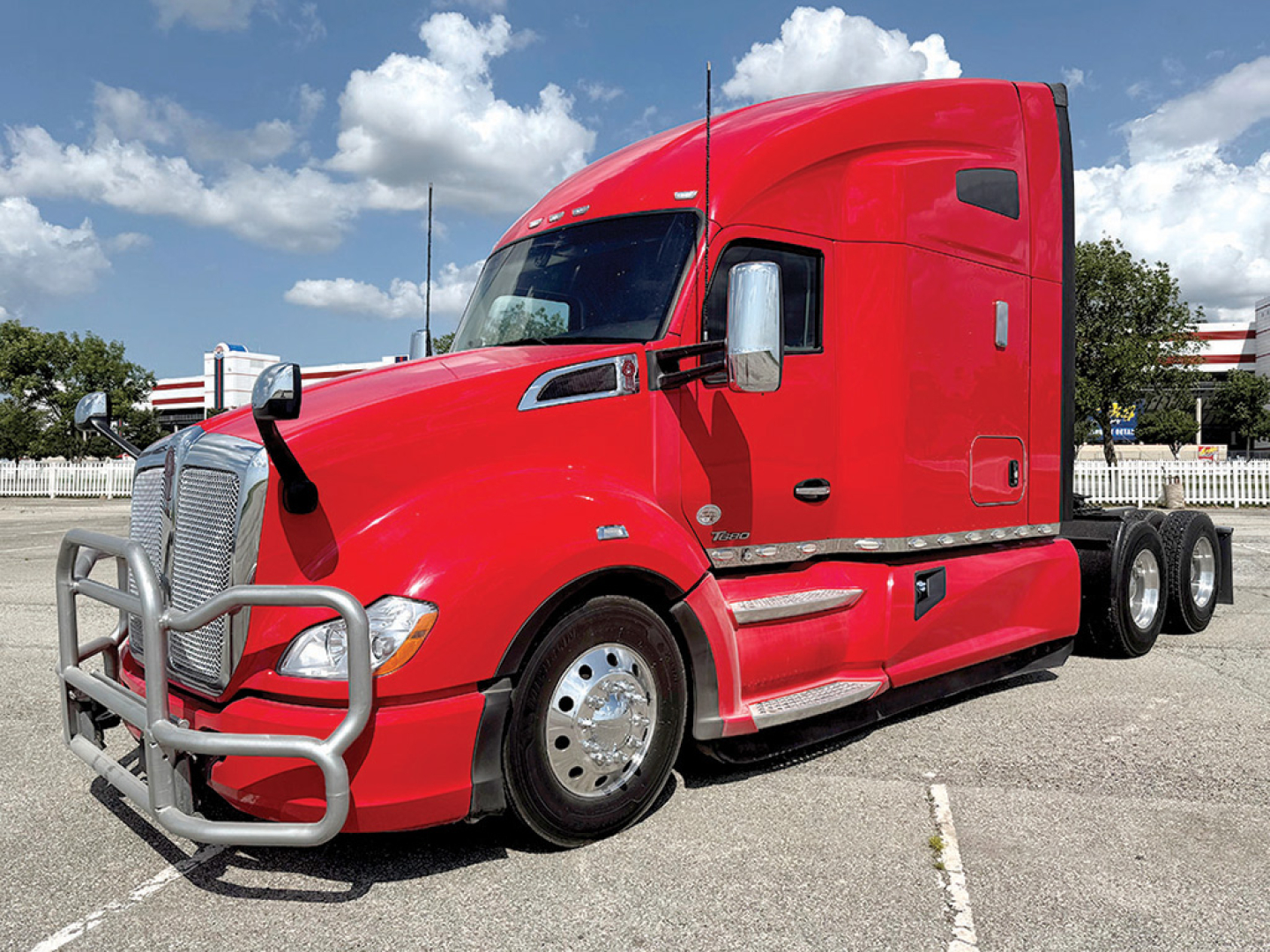 Kenworth T680 red truck in a dealership lot, front grille guard visible, two side mirrors, sleeper cab, bumper and chrome wheels.