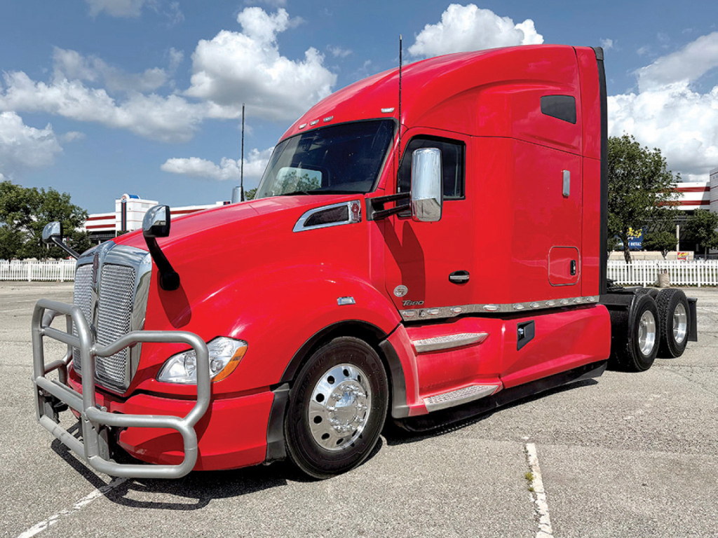 Kenworth T680 red truck in a dealership lot, front grille guard visible, two side mirrors, sleeper cab, bumper and chrome wheels.