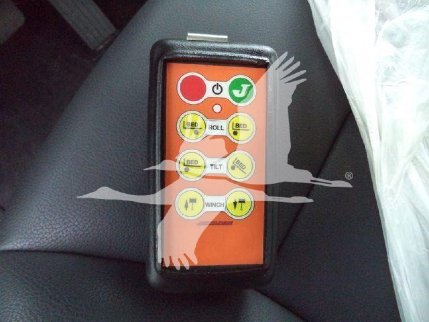 Remote control labeled with buttons: LIFT, ROLL, TILT, WINCH, PULL, plus symbols and icons; orange face with black border. Compact handheld control device.