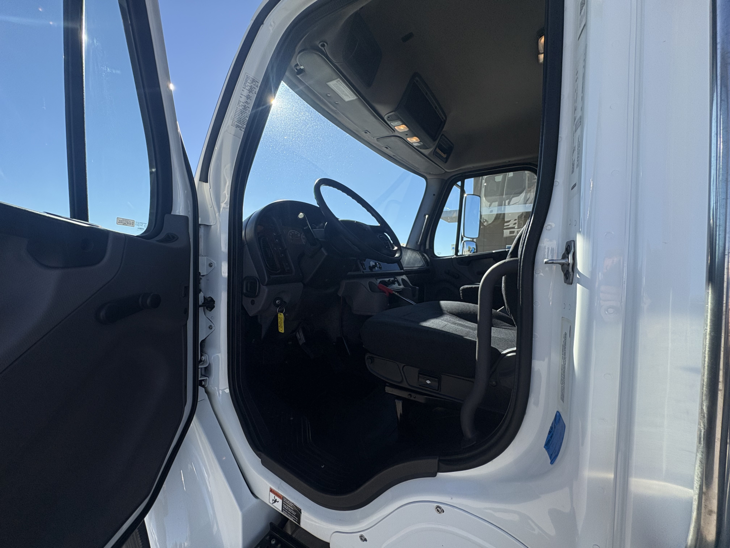 Freightliner M2-106 truck cab interior viewed from open door showing steering wheel, driver seat, pedals, and dashboard.