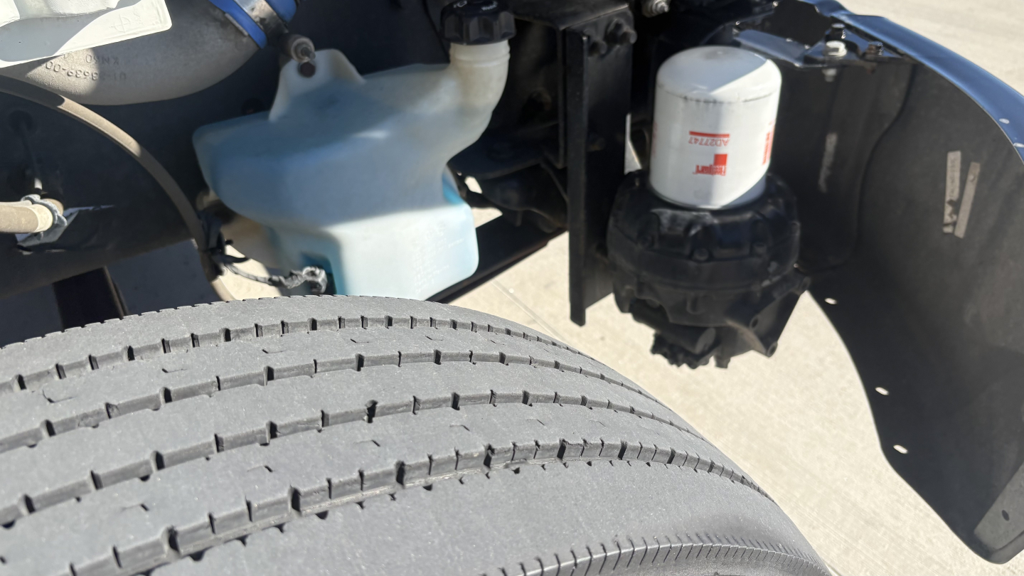 Freightliner M2-106 engine bay showing coolant reservoir, windshield washer bottle, and filter canister adjacent to a large front tire.