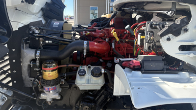 Engine bay of a Freightliner M2-106 with red engine components, hoses, and battery visible