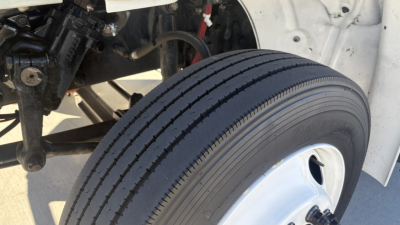 Truck wheel and suspension area; front left tire visible with white rim, undercarriage components, steering linkage, and part of white truck body.