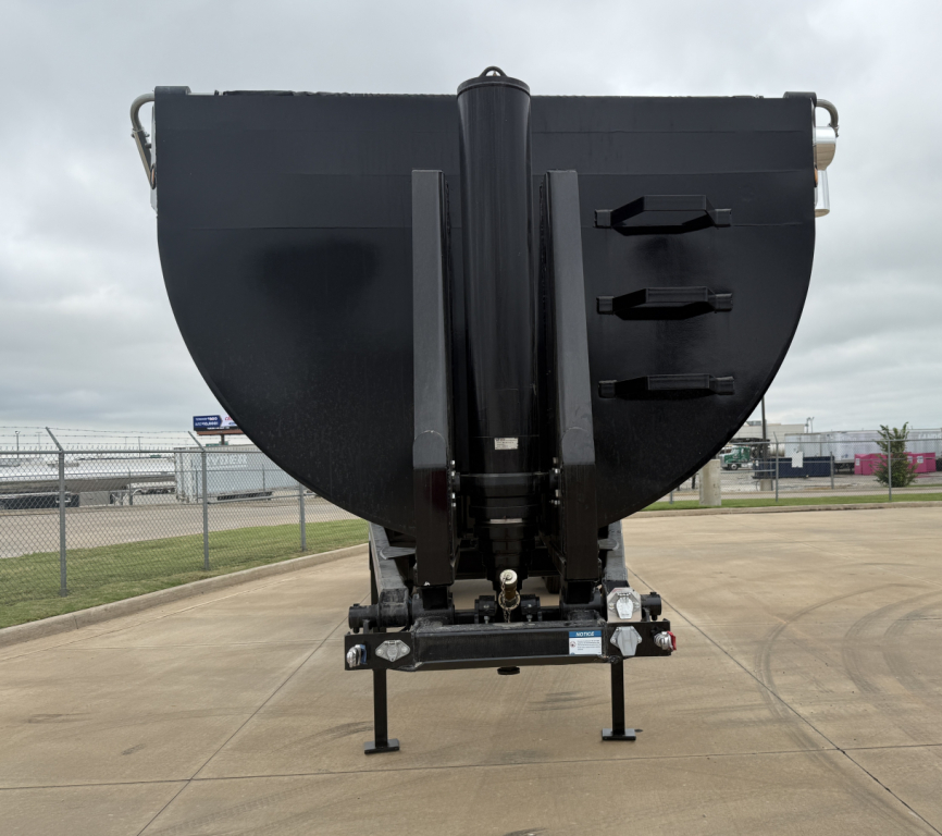Dump trailer in front view showing black armor and hydraulic lift with three tie-down steps on right side and front landing gear.