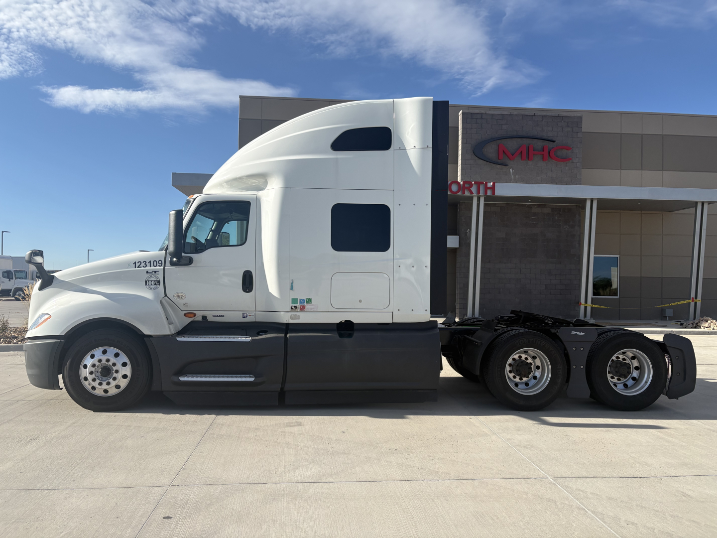 2023 International LT625 sleeper tractor side view showing class-7/8 tractor with white cab and black chassis, triple rear axles, and dealer building in background.